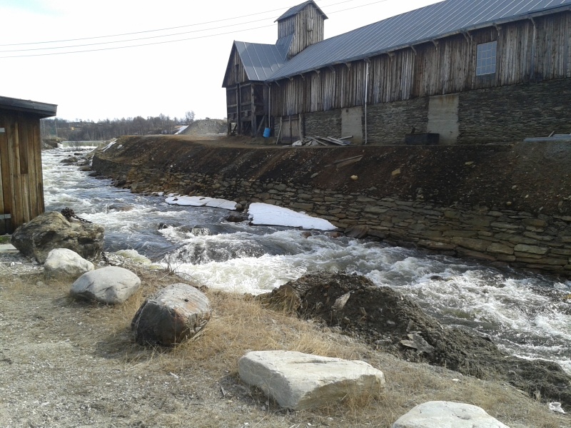 Gammal byggnad vid fors i Röros, i förgrunden grusparkering med stora stenar som avgränsning mot älvkanten. 7 maj roros